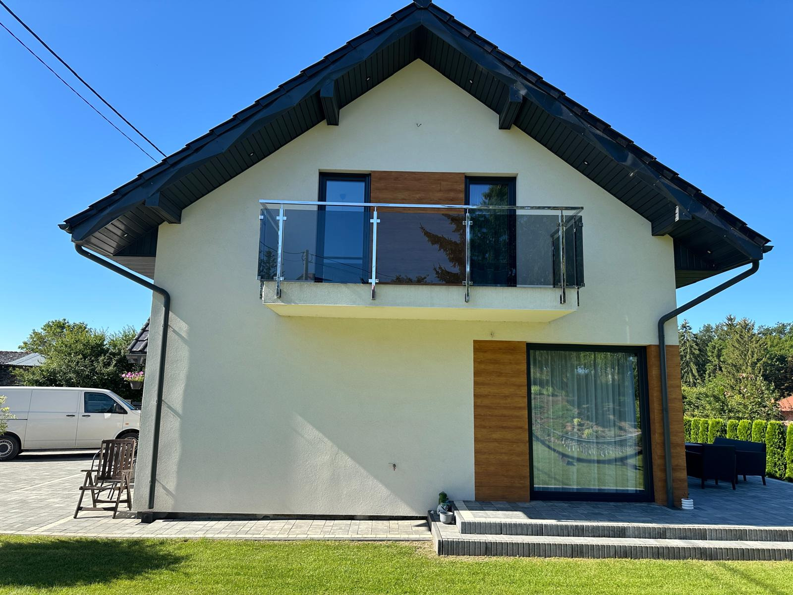 Nowoczesny dom z balkonem zabezpieczonym szklaną balustradą, widok z poziomu trawnika. Elementy wykończenia w odcieniach drewna, czarny dach i ramy okienne.