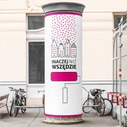 IMAGENCY Sp. z o.o. - Słup ogłoszeniowy z grafiką przedstawiającą stylizowane domy, różowe kropki imitujące deszcz, napis 'INACZEJ NIŻ WSZĘDZIE' i wałek do malowania, umieszczony na tle miejskiej architektury...