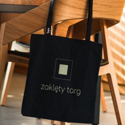 IMAGENCY Sp. z o.o. - Czarna torba bawełniana z nadrukiem logo i napisu 'zaklęty targ', zawieszona na drewnianym krześle.
