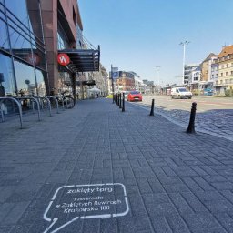 IMAGENCY Sp. z o.o. - Oznakowanie na chodniku w formie dymku z napisem 'zaklęty targ rzemiosło w Zaklętych Rewirach Krakowsko 100', widok na ulicę z budynkami i samochodami w tle, dzień.