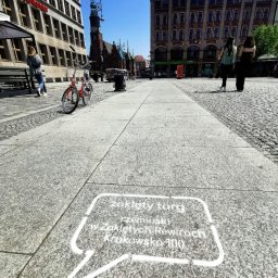 IMAGENCY Sp. z o.o. - Napis 'zaklety targ rzemiosło w Zakletych Rewirach Krakowskie 100' w ramce imitującej dymek na chodniku w centrum miasta, w tle budynki, rower i przechodnie.