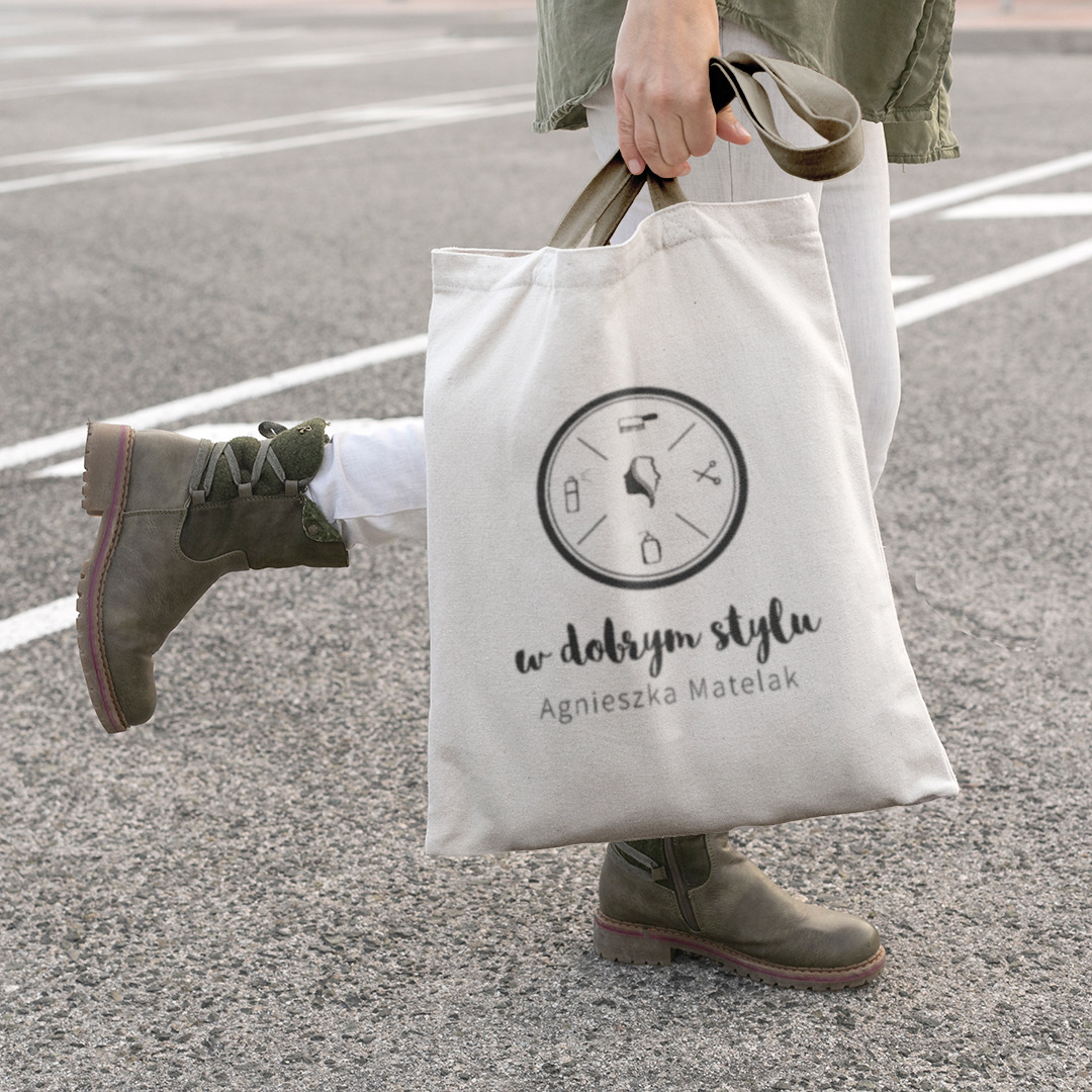 Osoba idąca z torbą bawełnianą z nadrukowanym logo i nazwą firmy 'w dobrym stylu Agnieszka Matelak', widoczne buty w kolorze khaki.