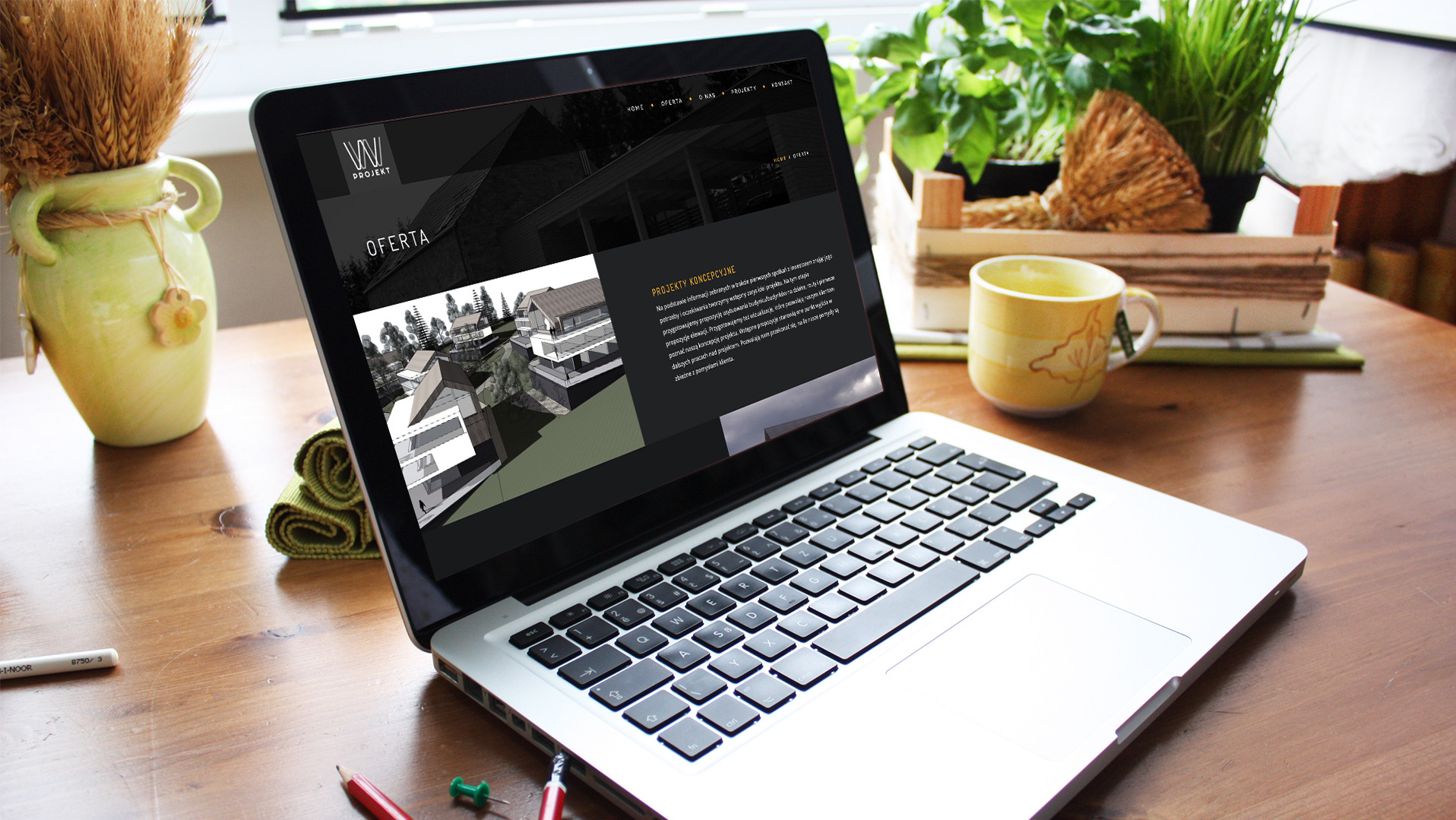 Laptop wyświetla stronę z wizualizacjami projektów architektonicznych, obok wazon z suszem, kubek i roślina doniczkowa na drewnianym biurku.
