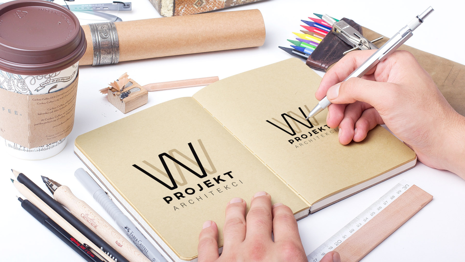 Ręka rysuje logo 'Projekt Architekci' w notatniku, obok przybory kreślarskie, kawa i ołówek