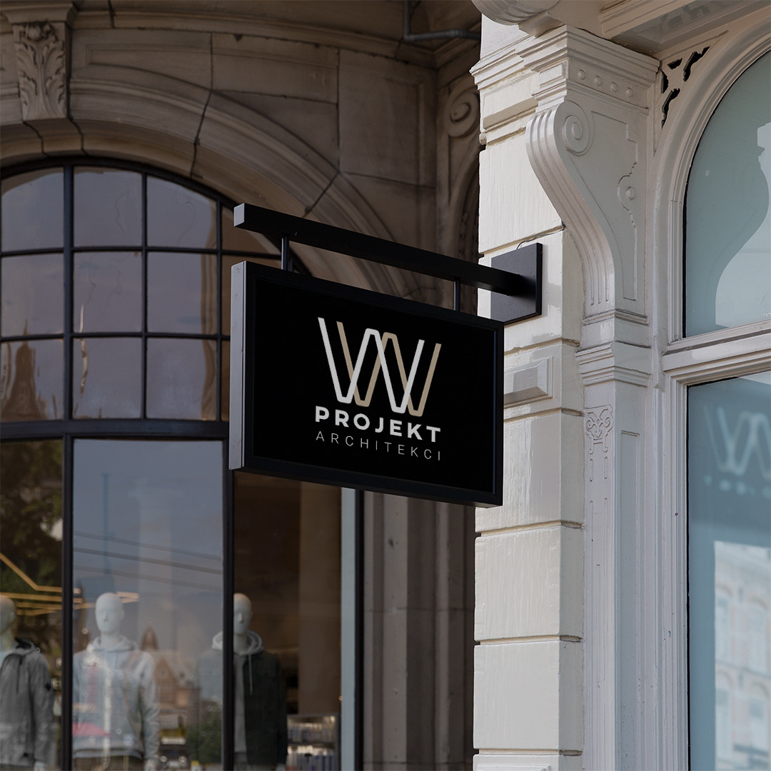 Szyld architektoniczny z logo 'Projekt Architekci' zawieszony na elewacji budynku z dużymi oknami wystawowymi.
