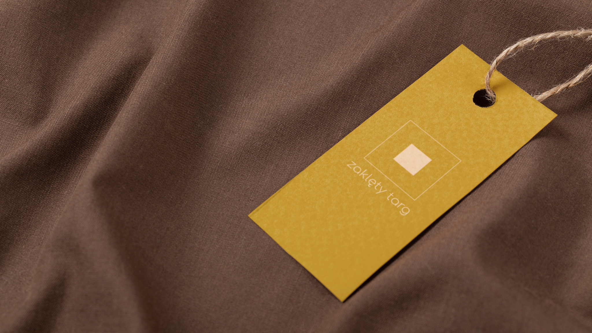 Żółta etykieta z logo i tekstem 'zaklęty targ' zawieszona na brązowej tkaninie z widoczną fakturą.
