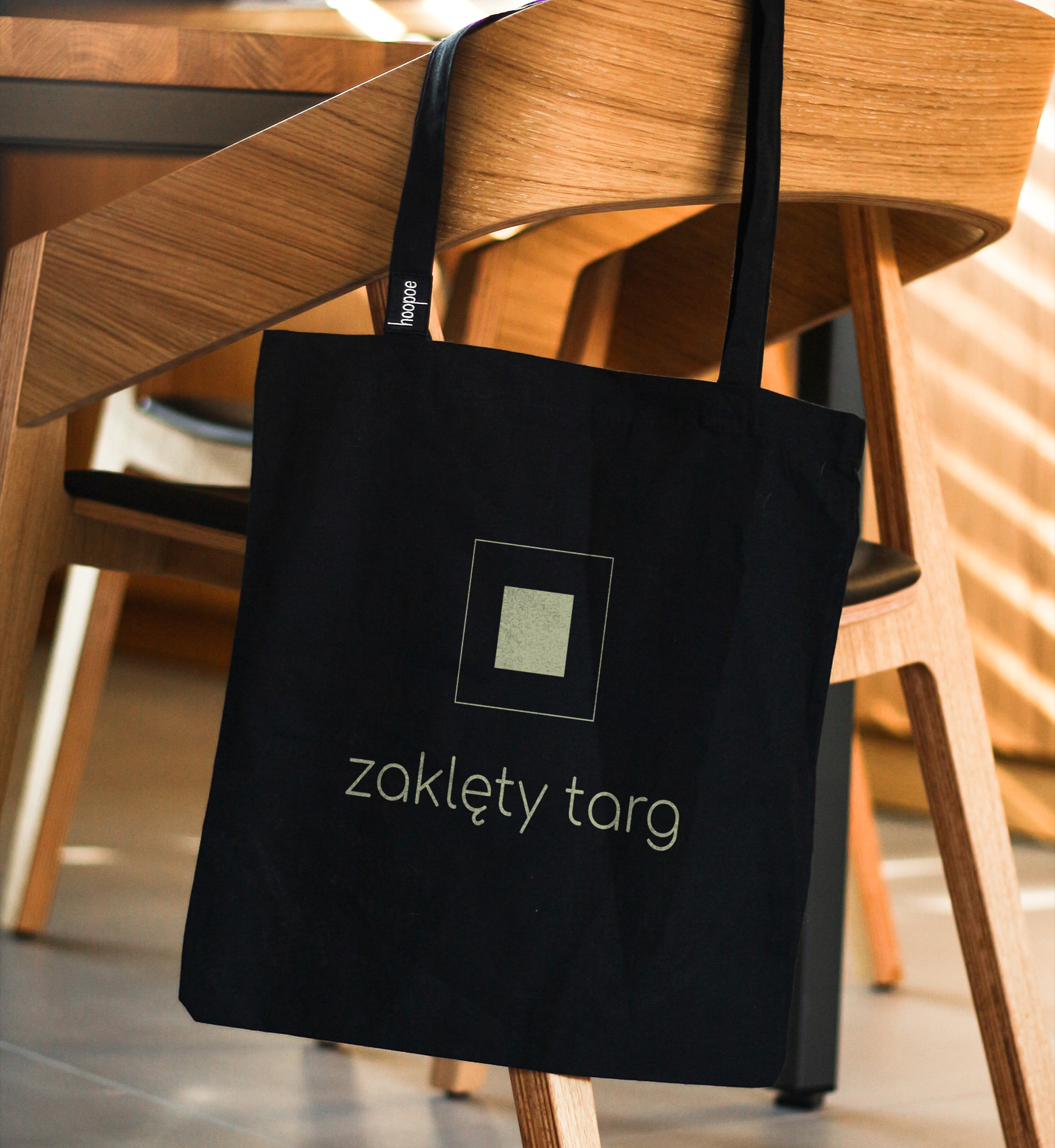 Czarna torba bawełniana z nadrukiem logo i napisu 'zaklęty targ', zawieszona na drewnianym krześle.