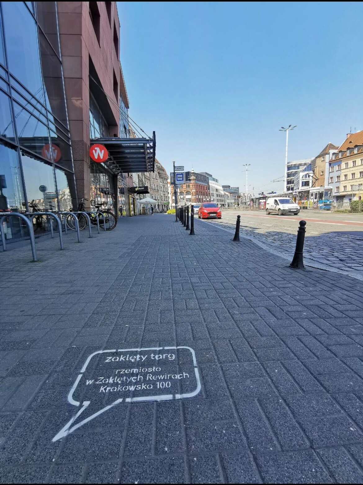 Oznakowanie na chodniku w formie dymku z napisem 'zaklęty targ rzemiosło w Zaklętych Rewirach Krakowsko 100', widok na ulicę z budynkami i samochodami w tle, dzień.