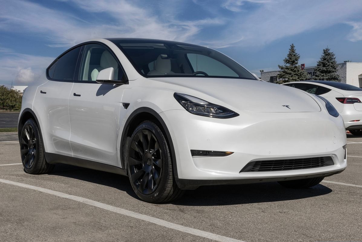 Biały samochód elektryczny Tesla Model Y z czarnymi felgami, zaparkowany na parkingu w słoneczny dzień, widoczny drugi samochód Tesla w tle.