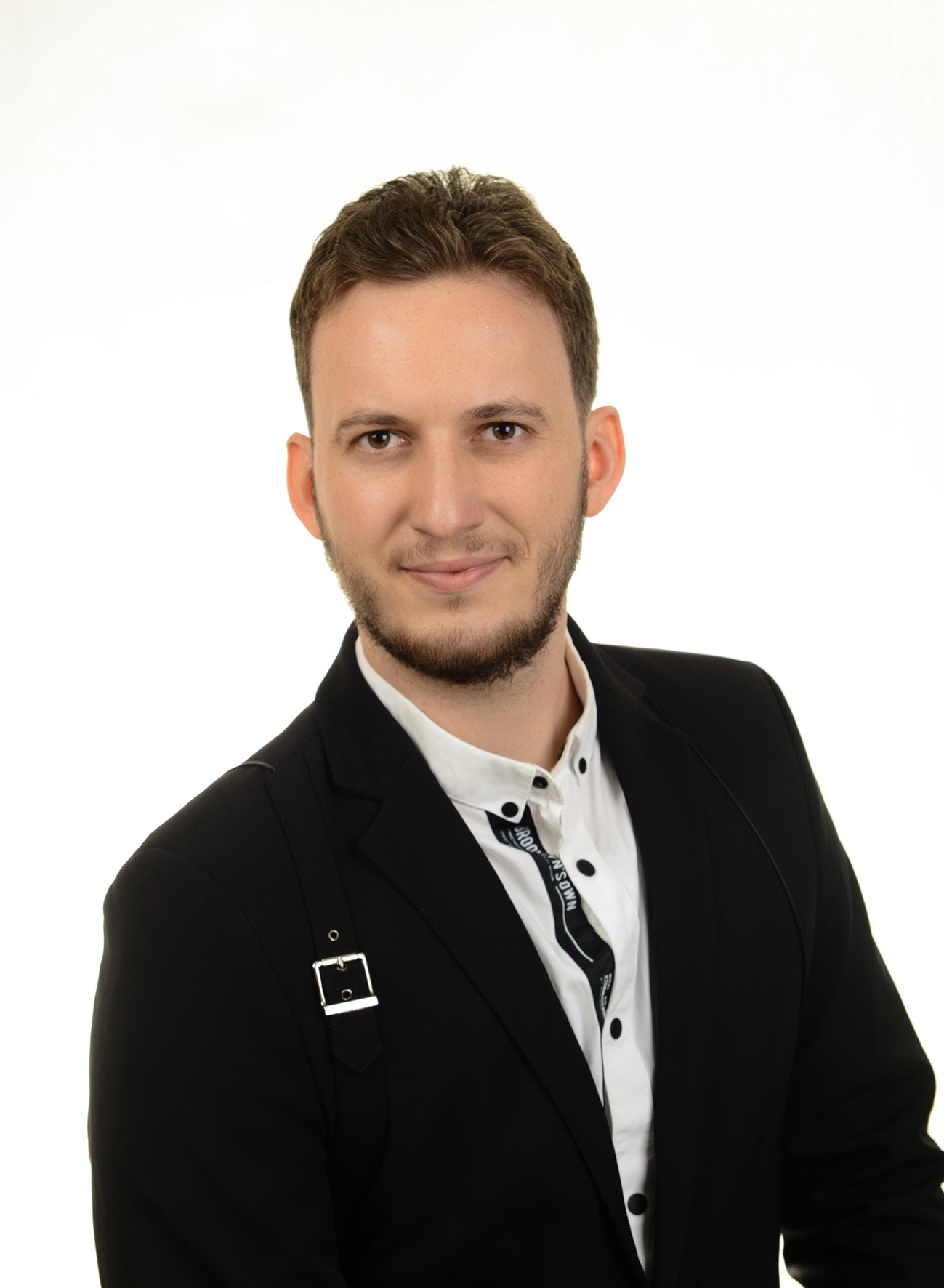 Portret uśmiechniętego mężczyzny w czarnym garniturze i białej koszuli z czarnymi guzikami, z identyfikatorem na szyi i ozdobnym paskiem z klamrą na ramieniu, na jasnym tle.