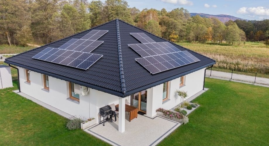 Nowoczesny dom jednorodzinny z białymi ścianami i ciemną dachówką, na której zamontowano panele fotowoltaiczne; widok z góry na zadbany trawnik, taras z grillem i otoczenie zalesione.