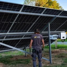 Montażysta w firmowej koszulce PCOZE stoi pod konstrukcją paneli fotowoltaicznych, widoczne okablowanie i inwerter, tło: drzewa i błękitne niebo.