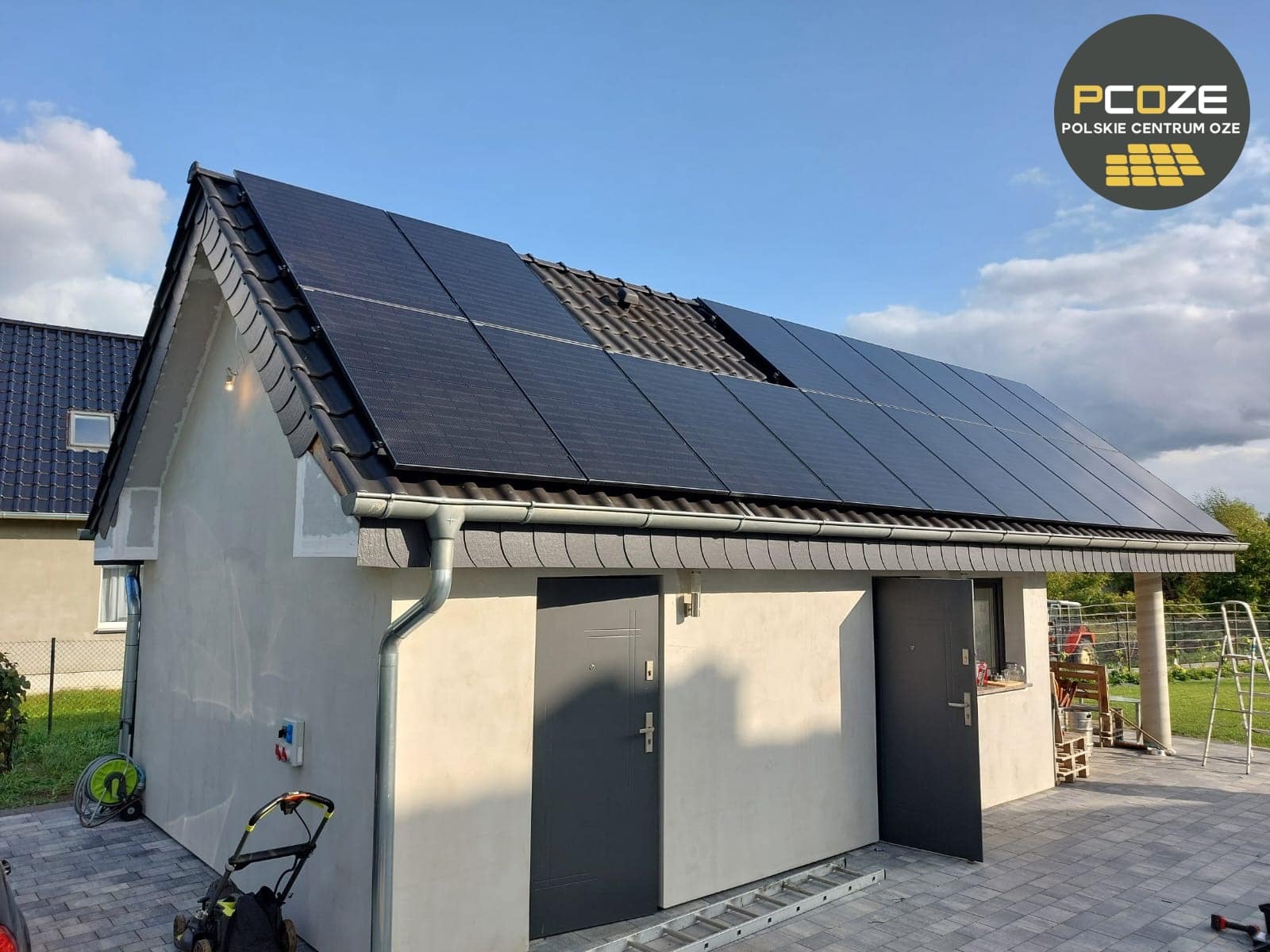 Instalacja paneli słonecznych na dachu budynku gospodarczego w Nysie, z widocznym fragmentem niezadaszonej połaci dachu i logiem firmy PC OZE w prawym górnym rogu.