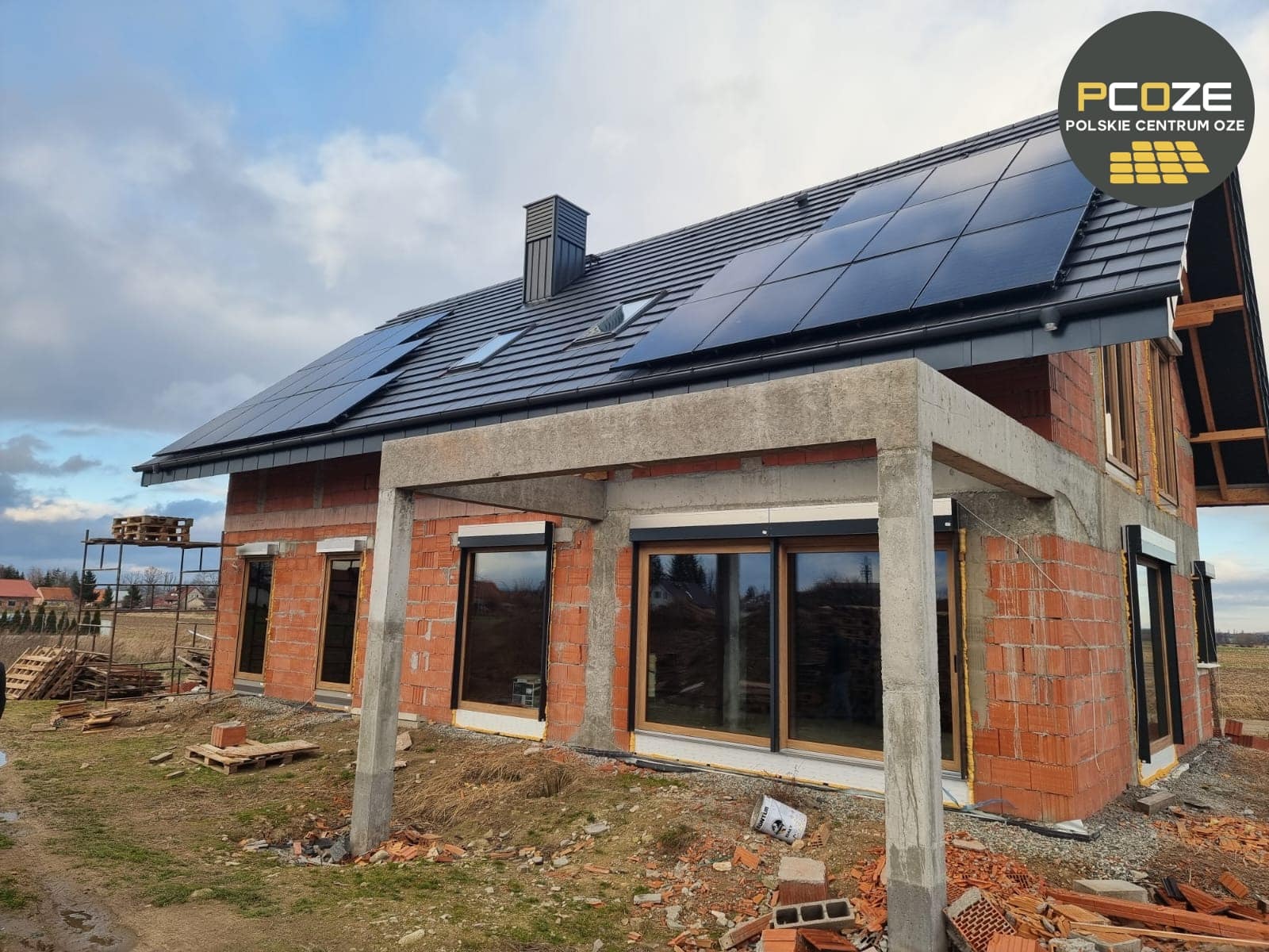 Niedokończony dom z czerwonej cegły z zamontowanymi panelami słonecznymi na dachu, widoczny logo firmy na górze po prawej stronie, okna zabezpieczone roletami zewnętrznymi, na zewnątrz porozrzucane...