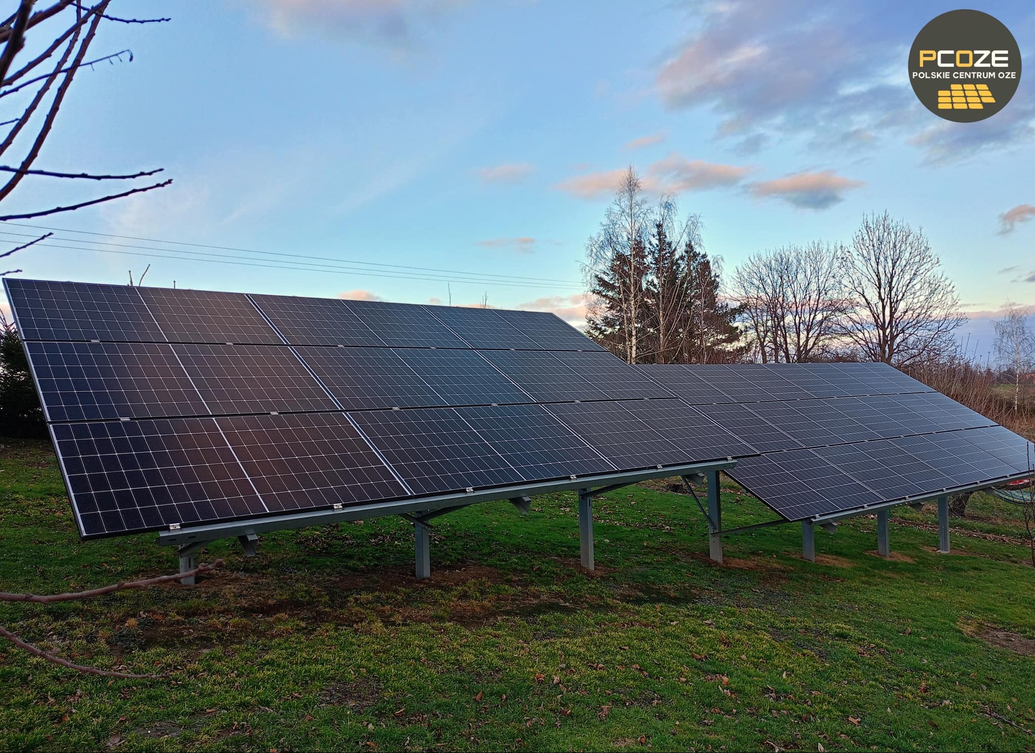 Dwa rzędy paneli słonecznych zamontowanych na metalowych konstrukcjach na trawiastym terenie, widoczne drzewa i błękitne niebo z chmurami w tle, w prawym górnym rogu logo firmy PCOZE.