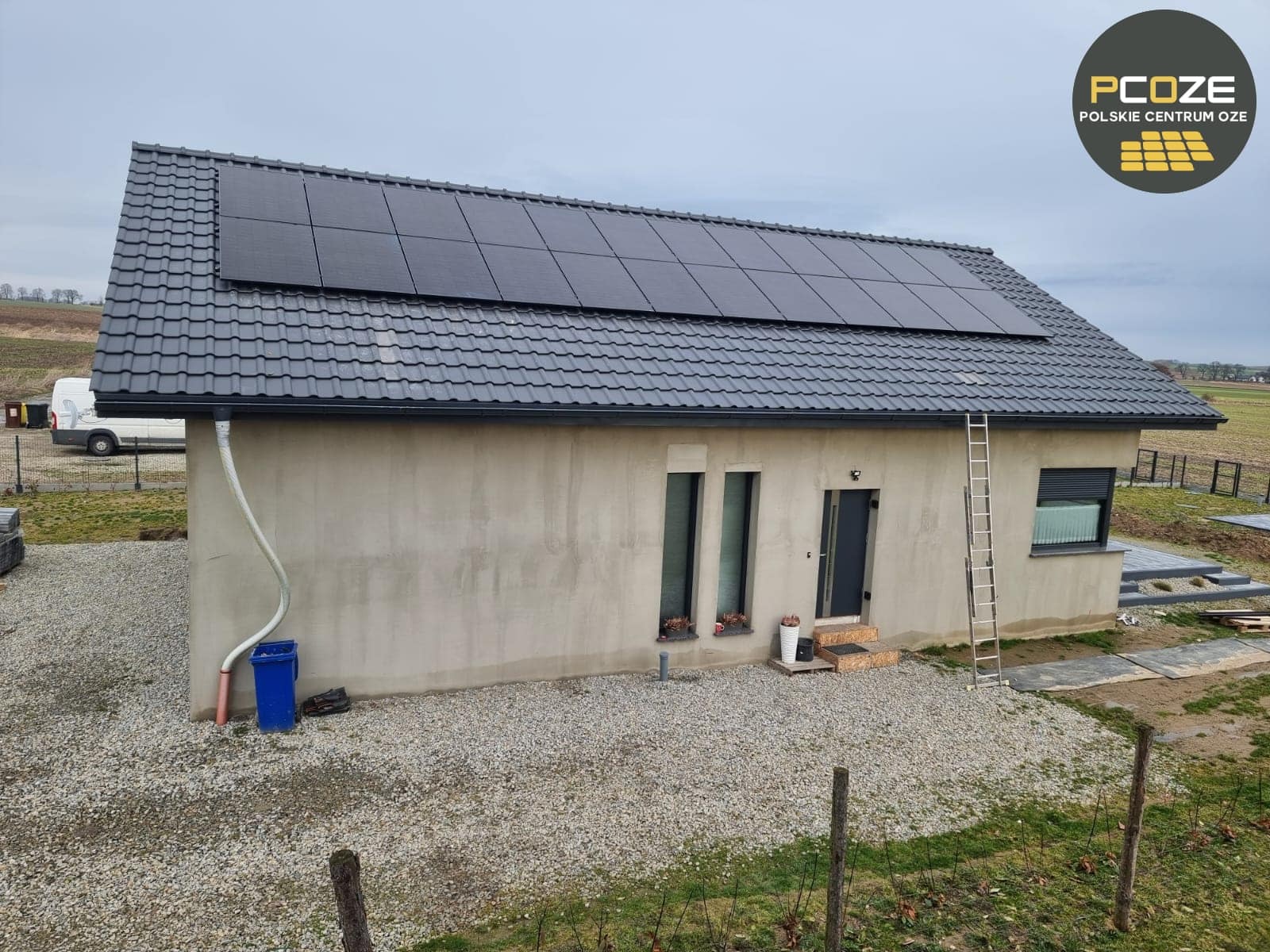 Dom jednorodzinny z szarym dachem, na którym zamontowano czarne panele słoneczne. Po lewej stronie widoczny niebieski pojemnik i biała rura. Po prawej drabina oparta o ścianę. W tle pola uprawne...