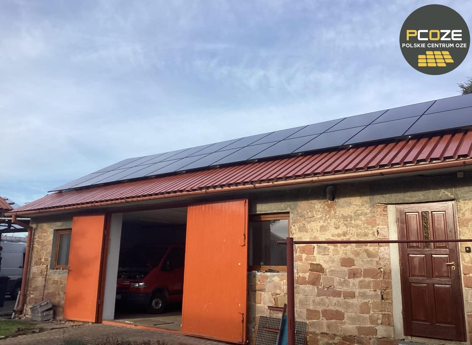 Instalacja paneli słonecznych na dachu budynku gospodarczego z czerwonej blachy trapezowej, widoczny otwarty garaż z czerwonym busem w środku oraz logo firmy w prawym górnym rogu.