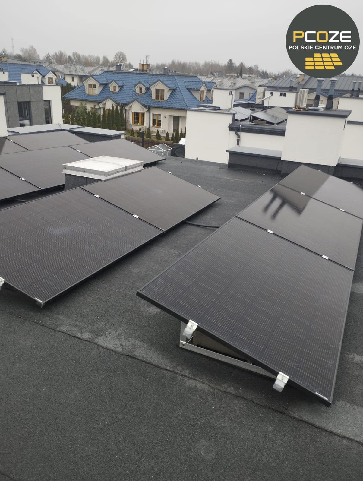 Panele fotowoltaiczne zamontowane na płaskim dachu budynku, widoczne metalowe uchwyty mocujące, w tle zabudowa jednorodzinna w pochmurny dzień, logo PCOZE w prawym górnym rogu.