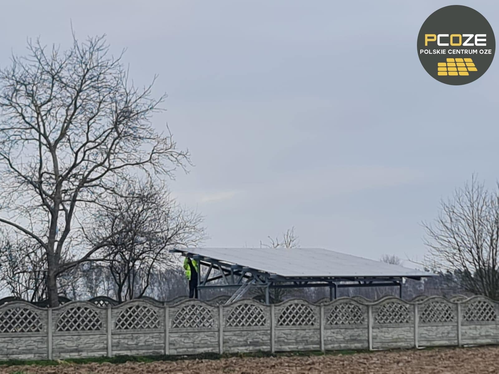 Montaż paneli słonecznych na konstrukcji wsporczej, widoczny pracownik w kamizelce odblaskowej, ogrodzenie betonowe i drzewa bez liści w tle, logo firmy w prawym górnym rogu.