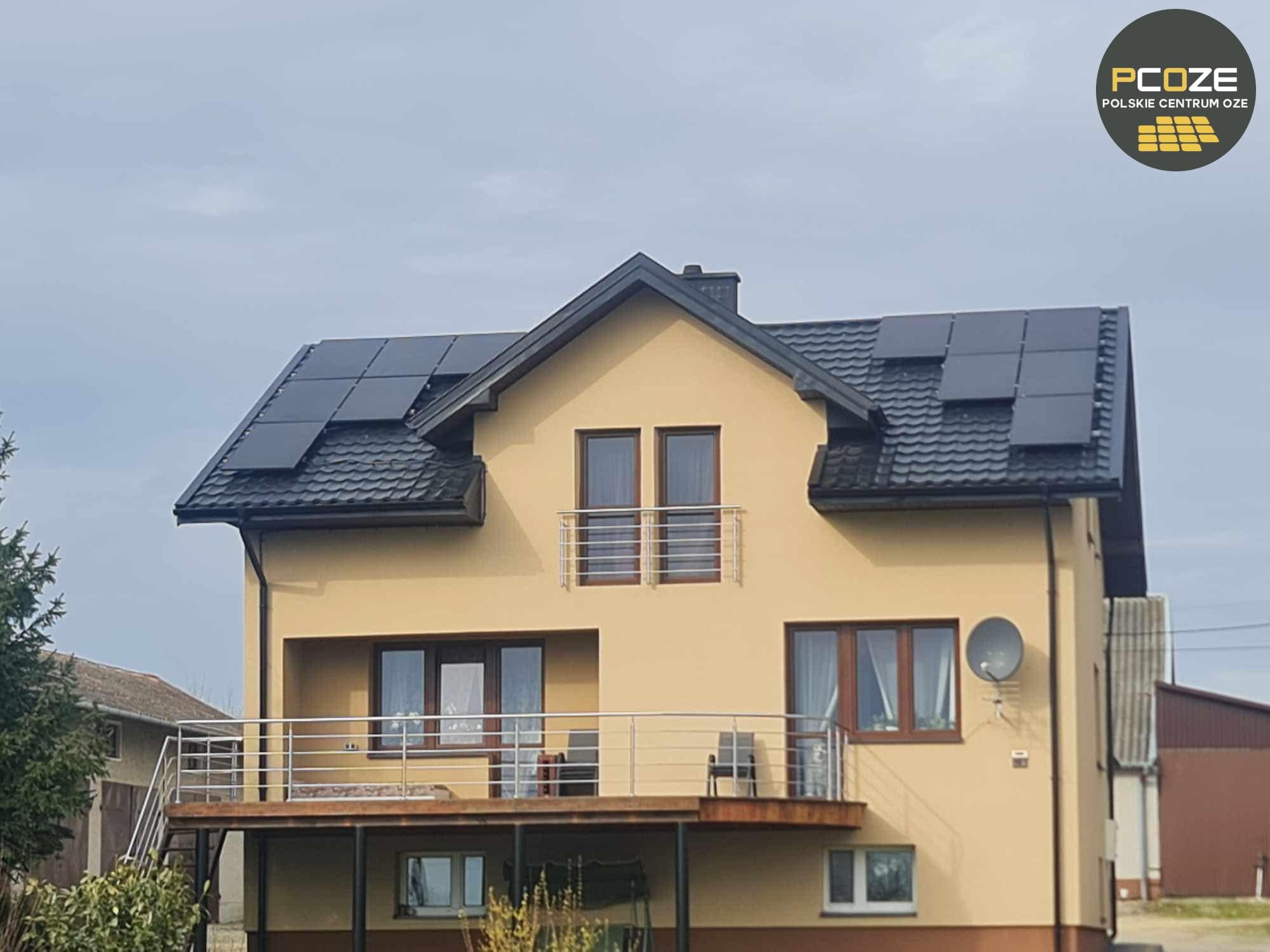Dwupiętrowy dom z beżową elewacją i czarnym dachem, na którym zamontowano panele słoneczne; balkon z metalową balustradą, logo firmy PCOZE widoczne w prawym górnym rogu.