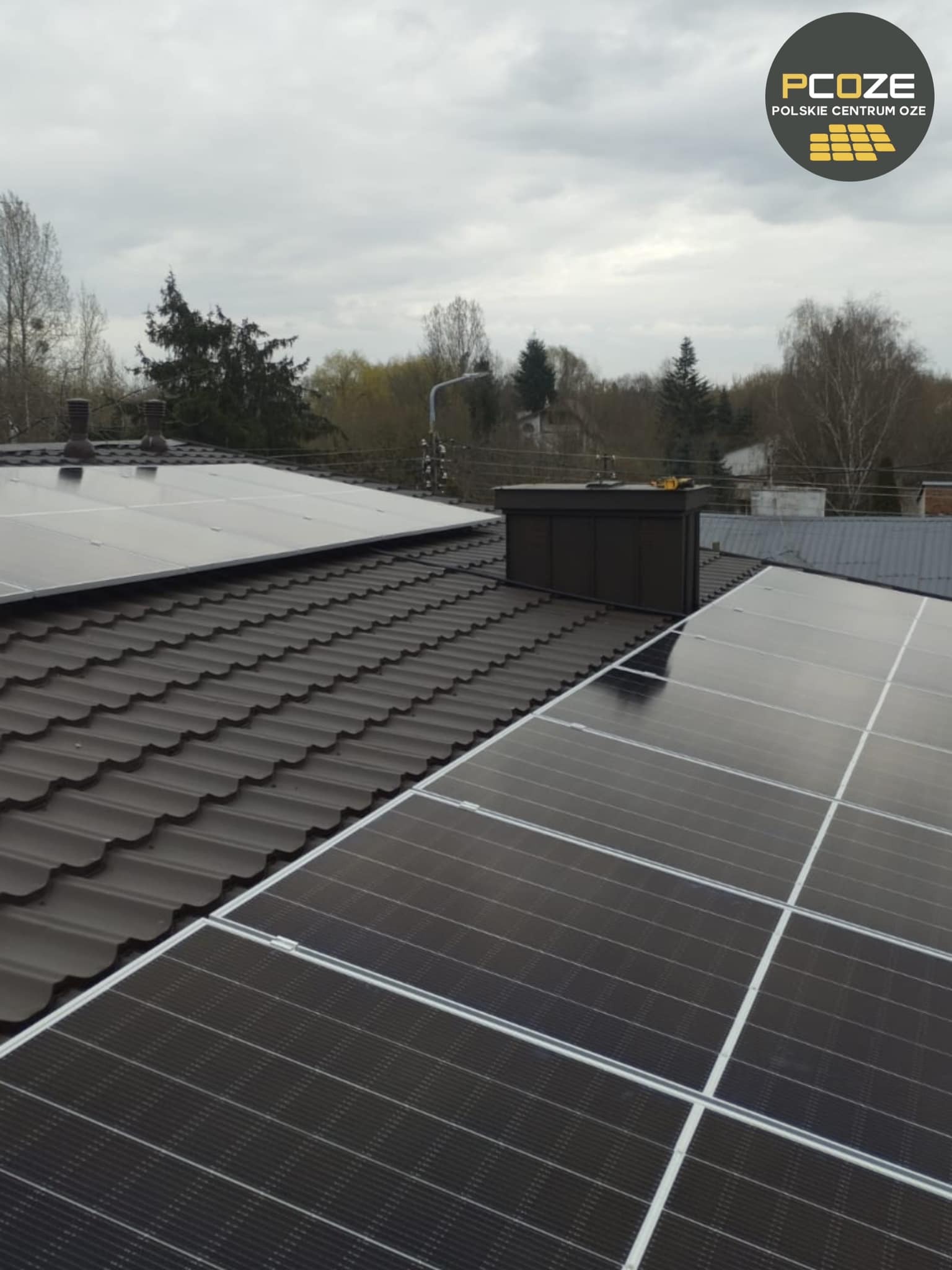 Panele słoneczne na ciemnobrązowym dachu z dachówki ceramicznej, widoczne kominy i logo firmy PCOZE w prawym górnym rogu, pochmurne niebo w tle.