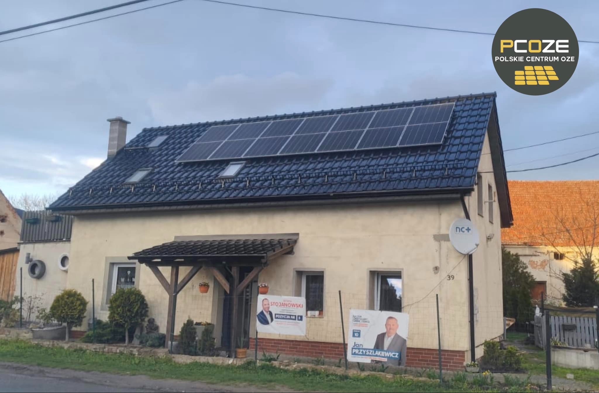 Dom jednorodzinny z zainstalowanymi panelami słonecznymi na ciemnym, spadzistym dachu, widoczne również okna dachowe, na elewacji budynku reklamy wyborcze polityków, a obok antena satelitarna.