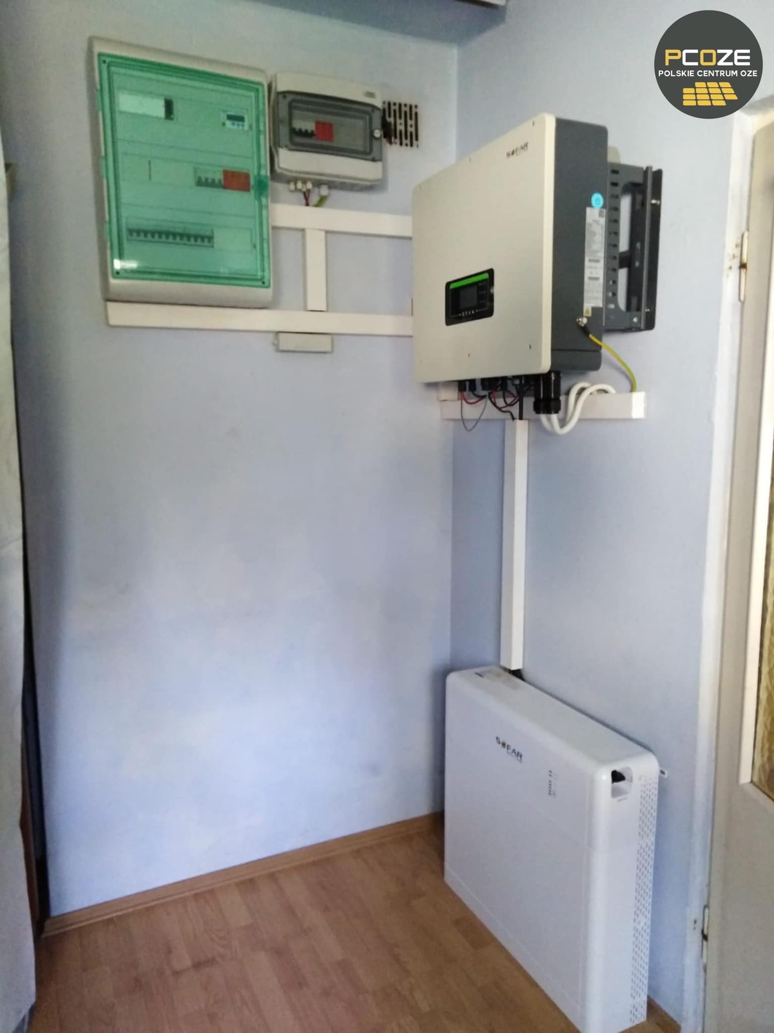Instalacja fotowoltaiczna wewnątrz budynku: rozdzielnica elektryczna, falownik z okablowaniem oraz akumulator na podłodze przy ścianie pomalowanej na niebiesko.