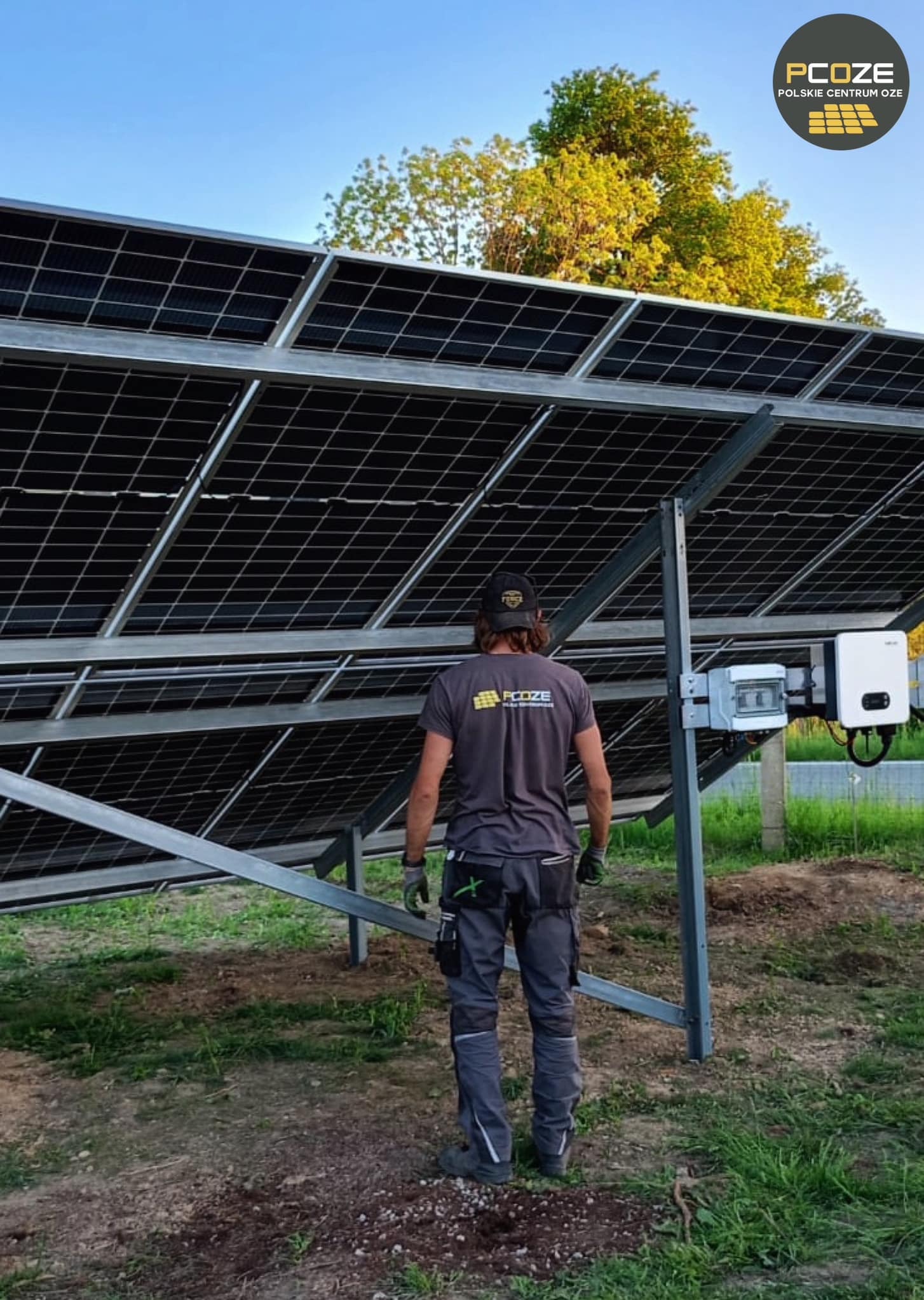 Montażysta w firmowej koszulce PCOZE stoi pod konstrukcją paneli fotowoltaicznych, widoczne okablowanie i inwerter, tło: drzewa i błękitne niebo.