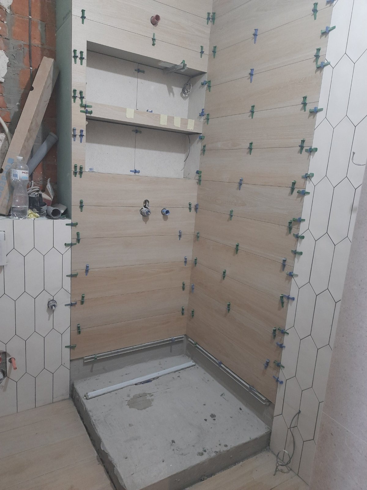 Łazienka w trakcie wykańczania: układanie płytek imitujących drewno na ścianach z użyciem klipsów poziomujących, widoczna wnęka z półkami i przygotowane podłoże pod prysznic.