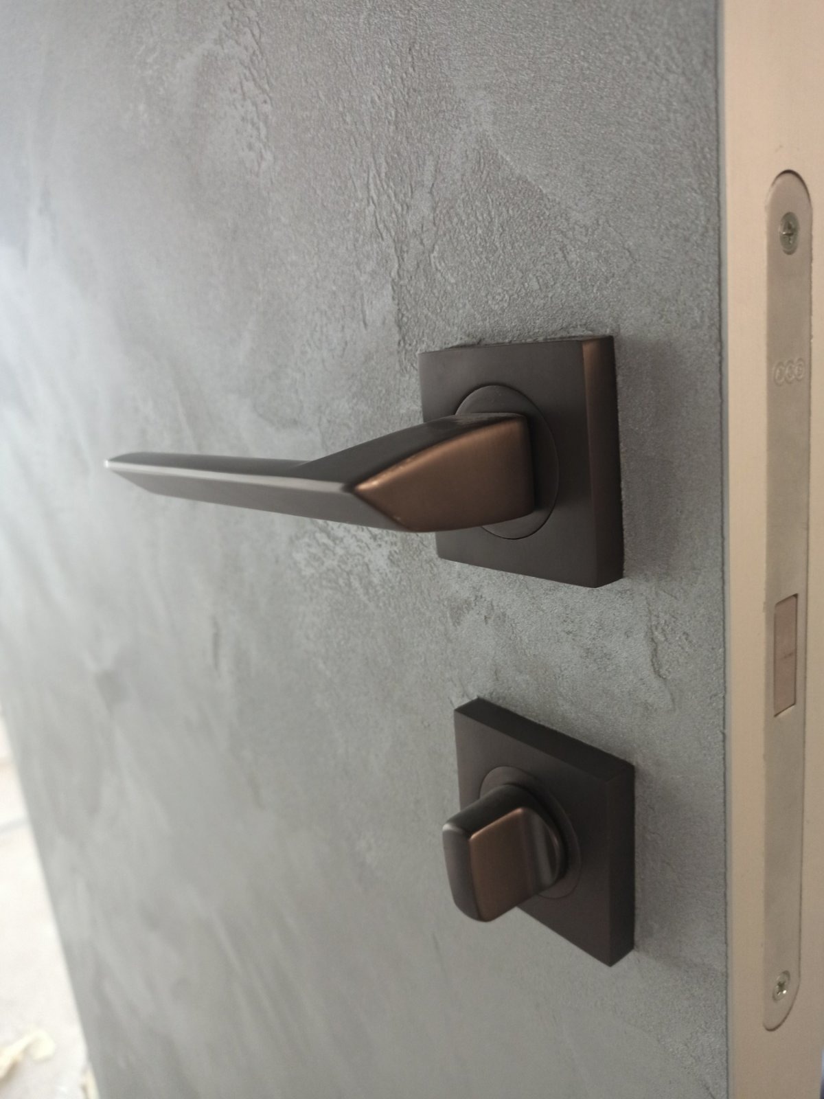 Szara ściana z dekoracyjną strukturą i nowoczesną, kwadratową klamką w odcieniach brązu, zamontowaną na białej ościeżnicy.