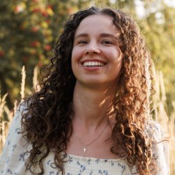 Kacper Pióro - Uśmiechnięta kobieta z kręconymi włosami w kwiecistej bluzce, pozująca na tle zieleni i wysokich traw w ciepłym, naturalnym świetle.