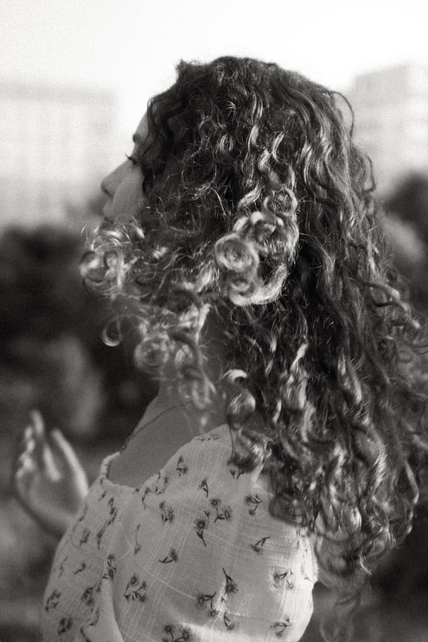 Czarno-białe zdjęcie profilu kobiety z kręconymi włosami, ubrana w bluzkę w drobne kwiatki, z delikatnym uśmiechem i dłońmi w tle.