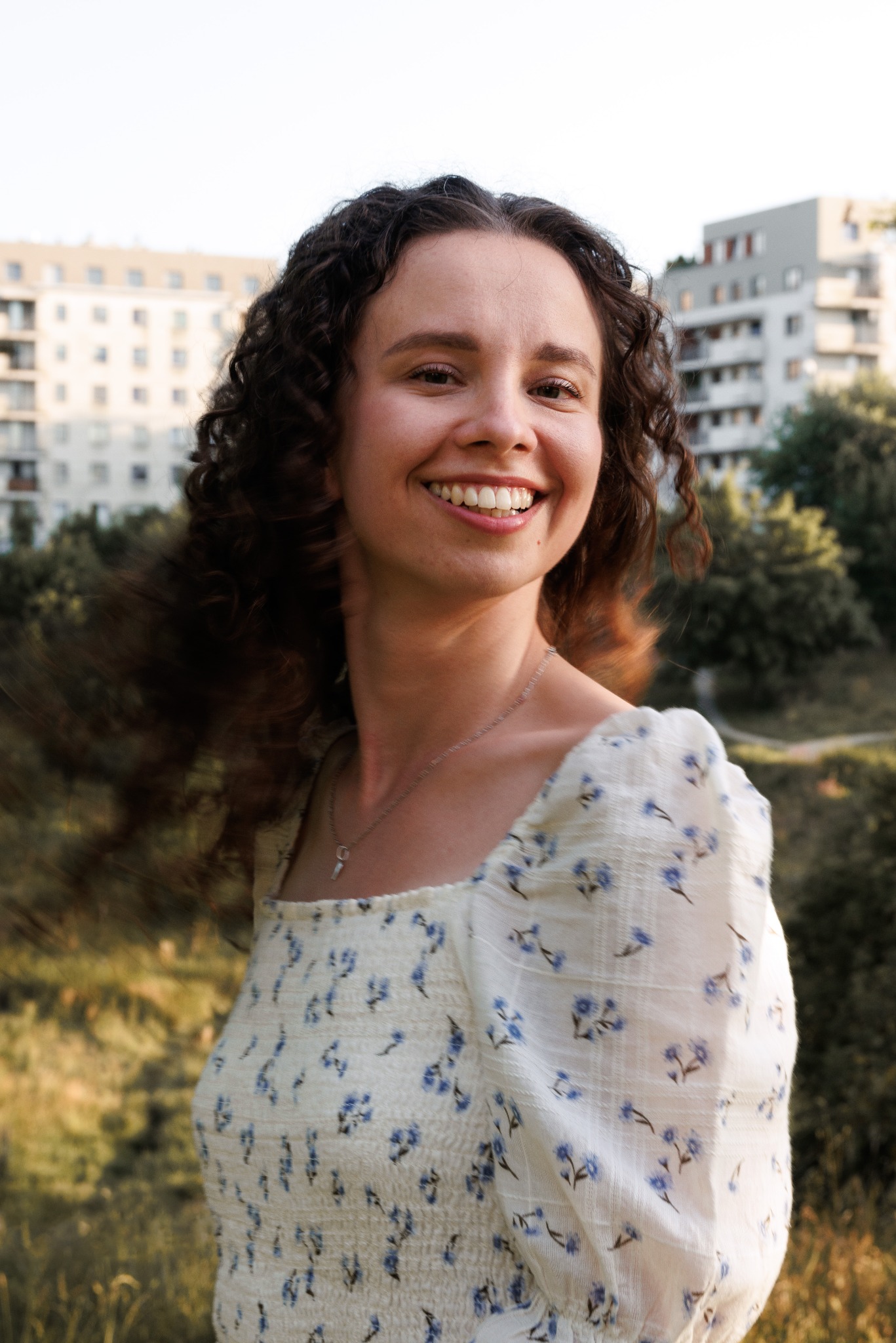 Uśmiechnięta młoda kobieta z kręconymi włosami w kwiecistej sukience, pozująca na tle zieleni i budynków mieszkalnych w ciepłym świetle słonecznym.