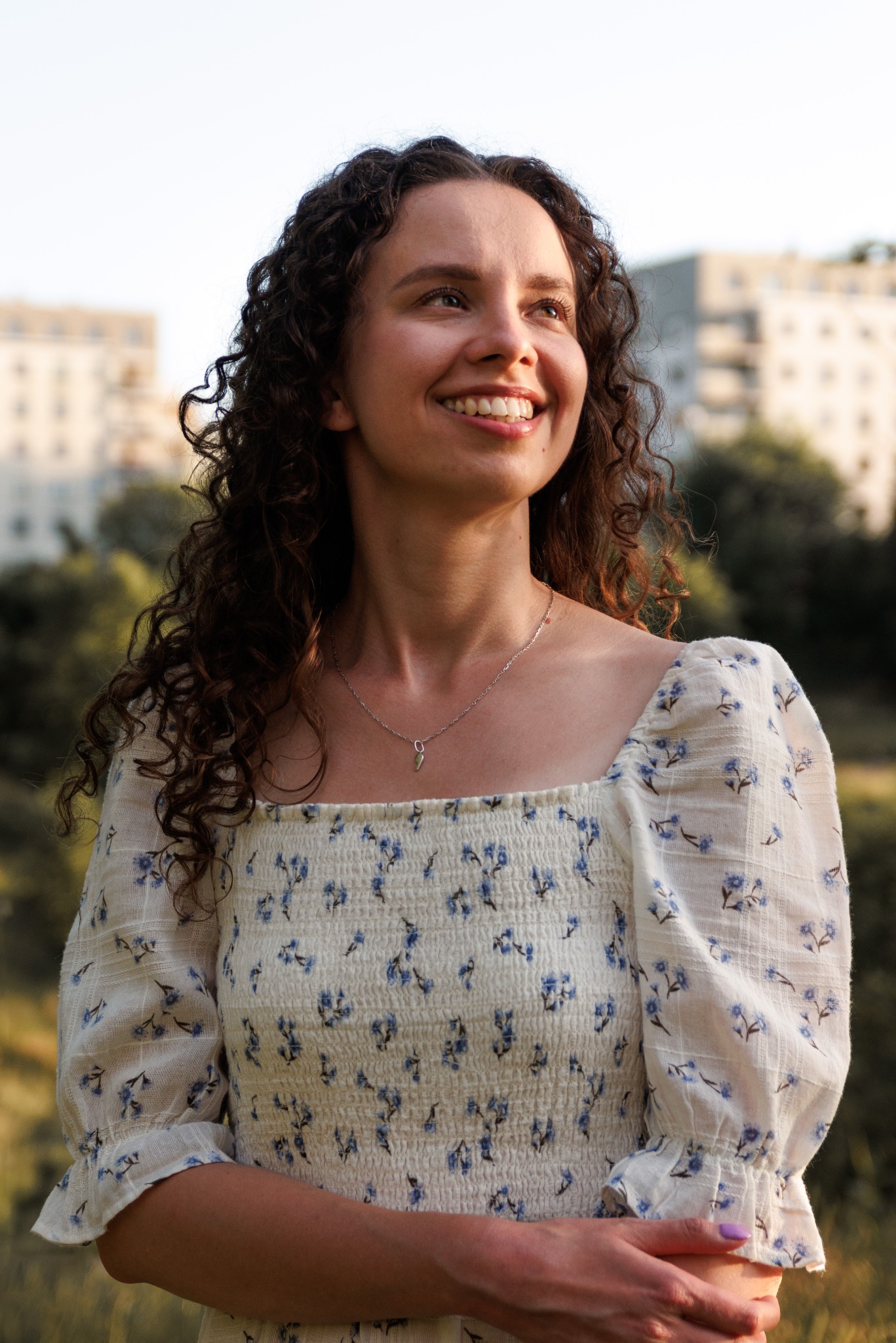 Portret uśmiechniętej kobiety z kręconymi włosami w białej sukience w drobne niebieskie kwiatki, na tle zieleni i budynków.