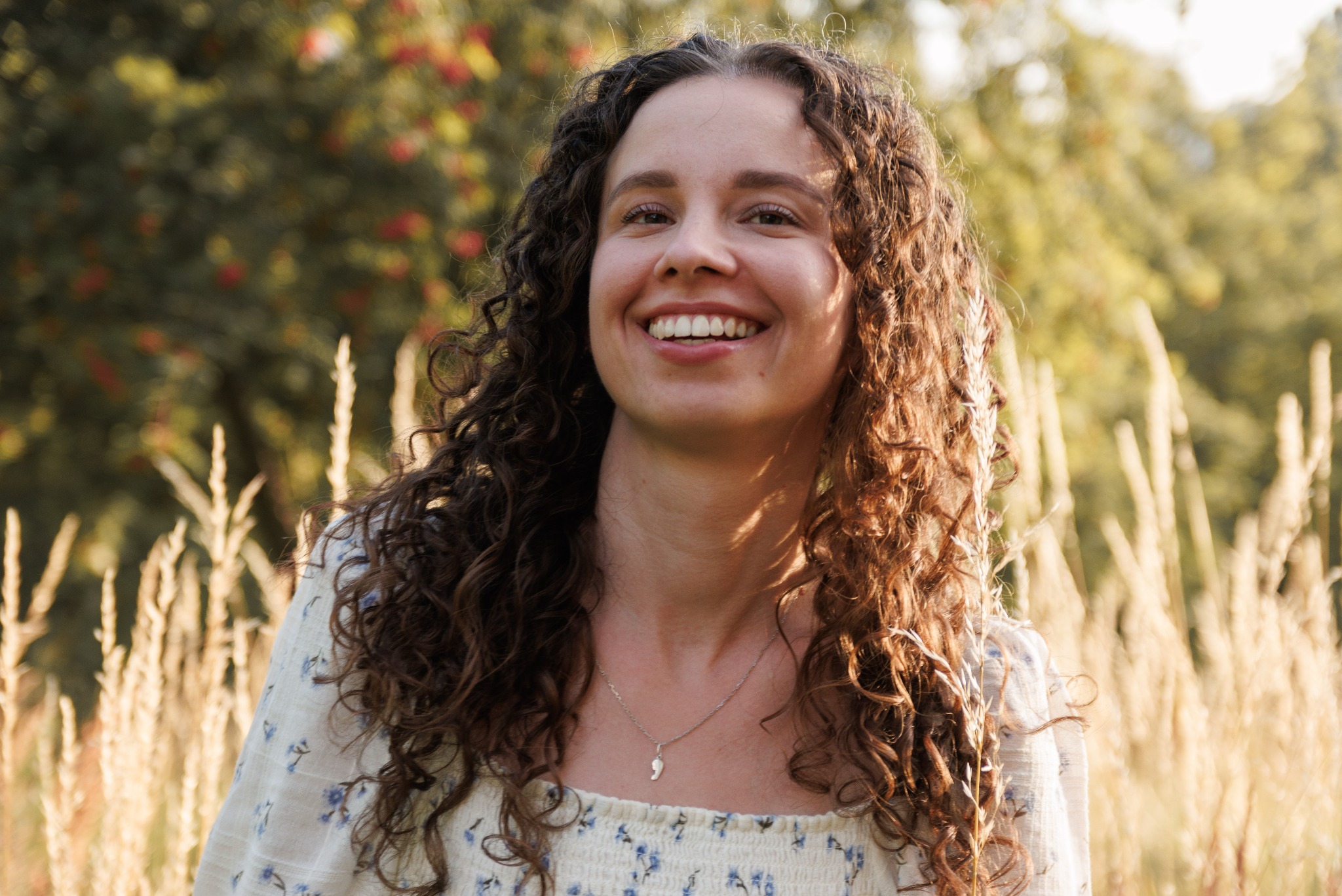 Uśmiechnięta kobieta z kręconymi włosami w kwiecistej bluzce, pozująca na tle zieleni i wysokich traw w ciepłym, naturalnym świetle.