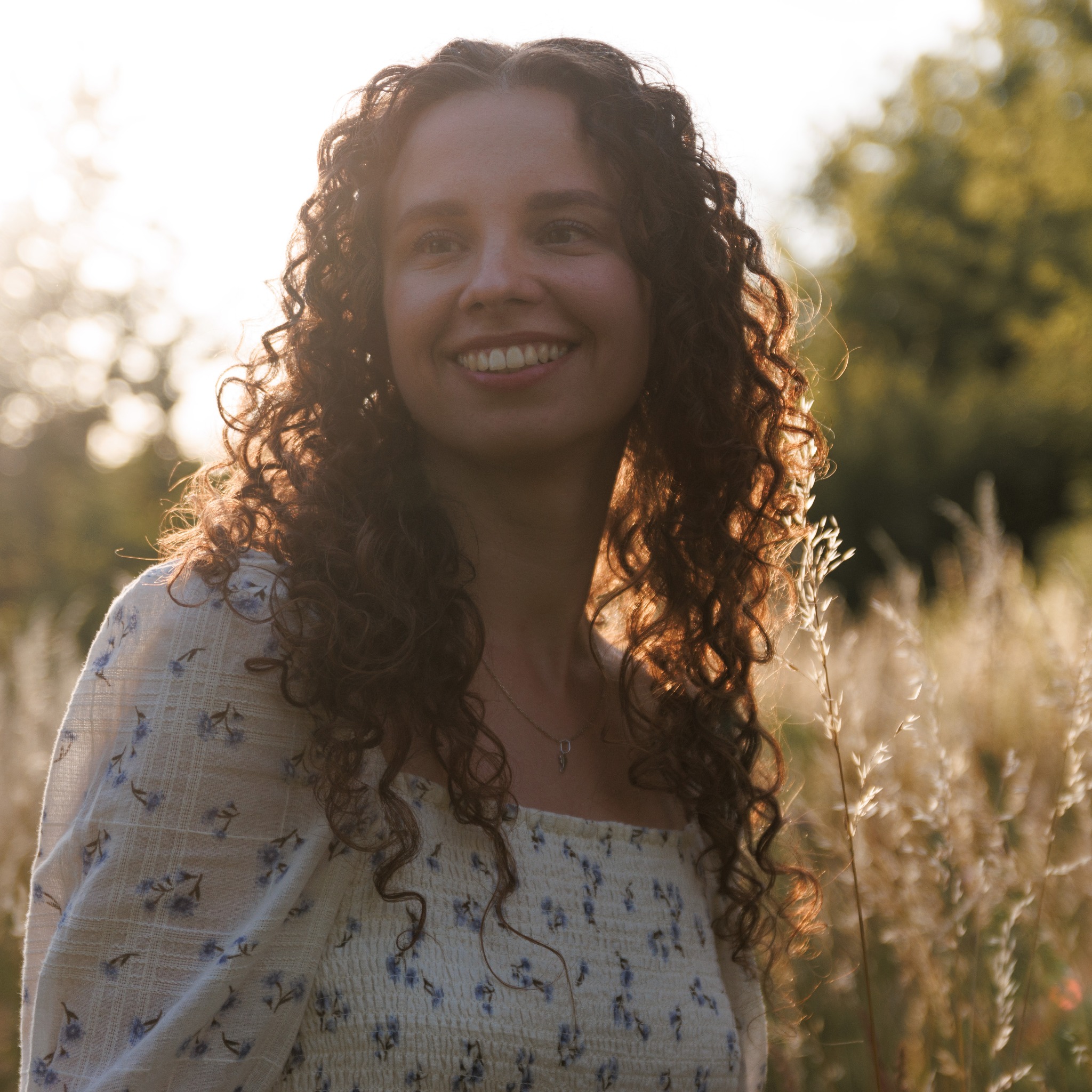 Portret uśmiechniętej kobiety z burzą kręconych włosów, oświetlonej zachodzącym słońcem w plenerze, na tle wysokiej trawy.