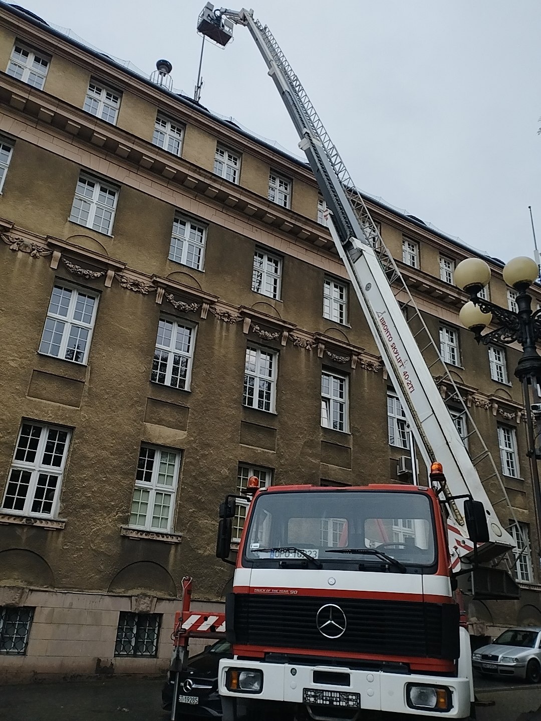 Czerwono-biały samochód z podnośnikiem koszowym na tle starej kamienicy podczas pracy na wysokości dachu.