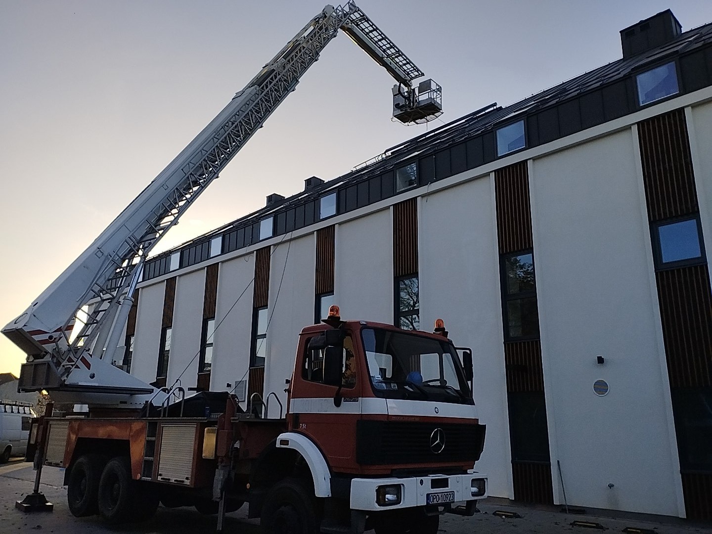 Ciężki samochód z wysuwanym ramieniem podnośnika koszowego ustawiony przed nowoczesnym budynkiem z białymi ścianami i ciemnymi wstawkami, prawdopodobnie w trakcie prac na wysokości.