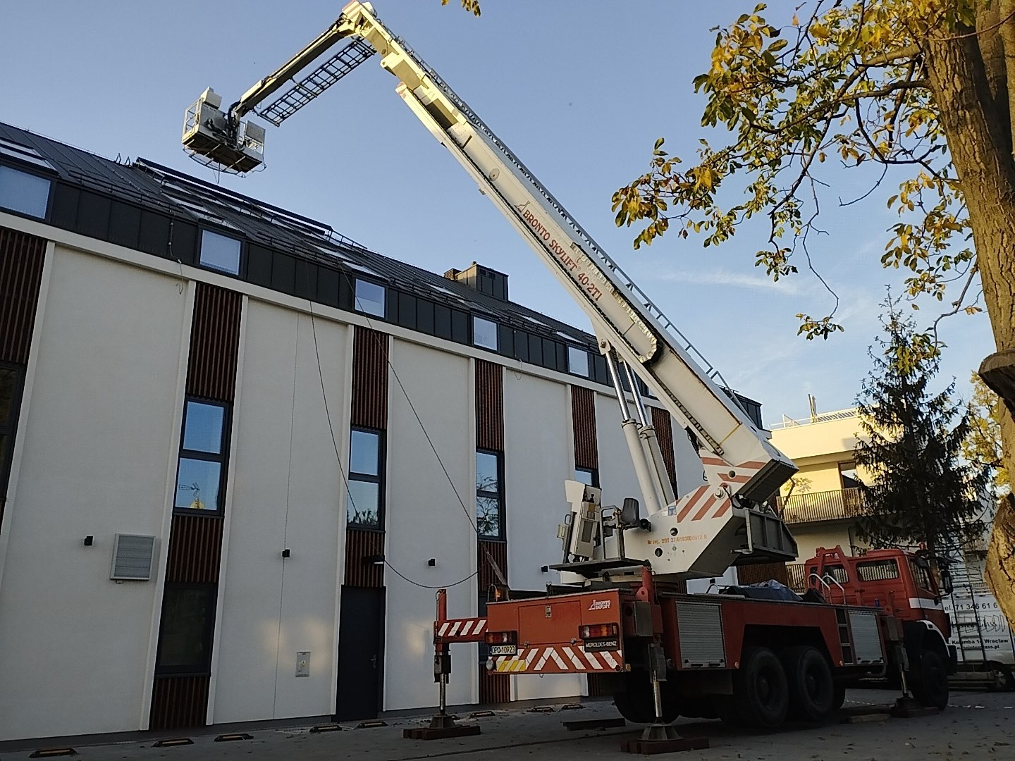 Czerwono-biały podnośnik koszowy Bronto Skylift 40-2T1 ustawiony przed nowoczesnym budynkiem z białymi ścianami i ciemnymi, pionowymi panelami dekoracyjnymi oraz czarnym dachem, kosz podnośnika...