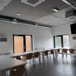 Wnętrze biura z systemem wentylacji: widoczne kanały wentylacyjne na suficie, kratki wentylacyjne w suficie podwieszanym, białe ściany, długi stół konferencyjny z krzesłami, szare panele podłogowe.