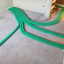 Zielone, karbowane rury wentylacyjne rozprowadzone po surowej podłodze w trakcie instalacji, zabezpieczone metalowymi opaskami i pianką montażową przy ścianach.