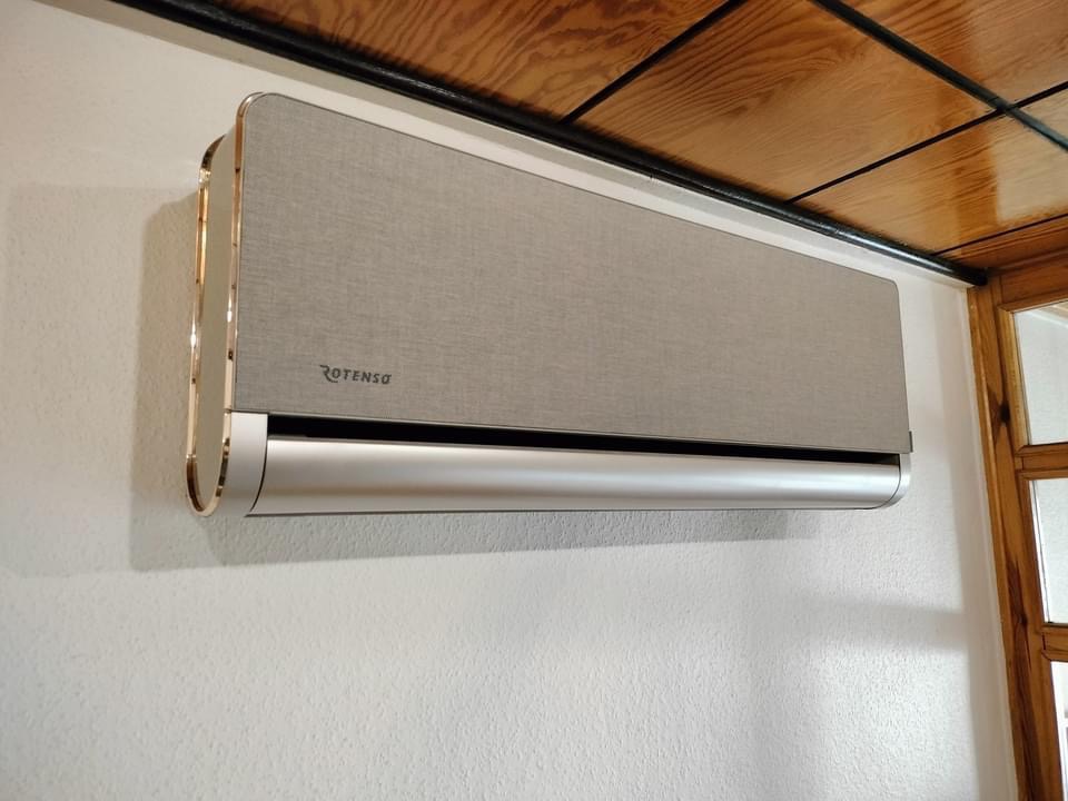 Szary klimatyzator Rotenso z chromowanymi wykończeniami, zamontowany na białej ścianie w pomieszczeniu z drewnianym sufitem i ościeżnicą.