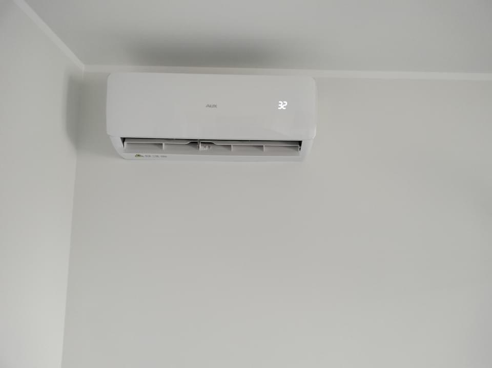 Biały klimatyzator marki AUX zamontowany na jasnej ścianie w rogu pokoju, wyświetlający temperaturę 32 stopnie.
