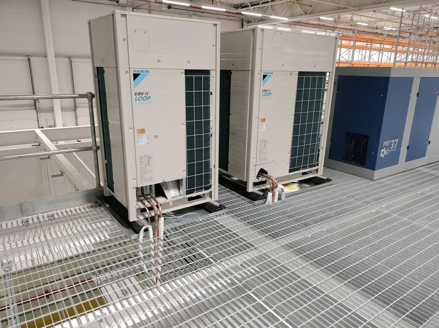 Dwa moduły klimatyzacji przemysłowej Daikin VRV IV Loop ustawione na metalowej platformie w hali produkcyjnej, z widocznymi rurami miedzianymi i niebieską sprężarką w tle.