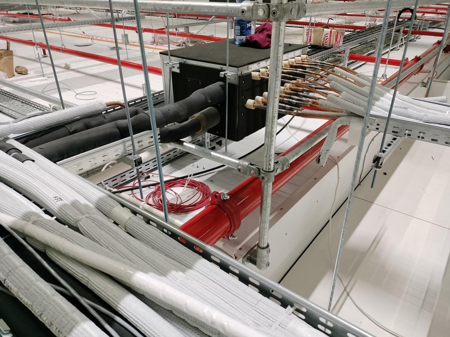 Montaż instalacji rekuperacji w budynku komercyjnym: widok na centralę rekuperacyjną z podłączonymi przewodami, rury w izolacji termicznej, czerwona rura instalacji przeciwpożarowej i elementy...