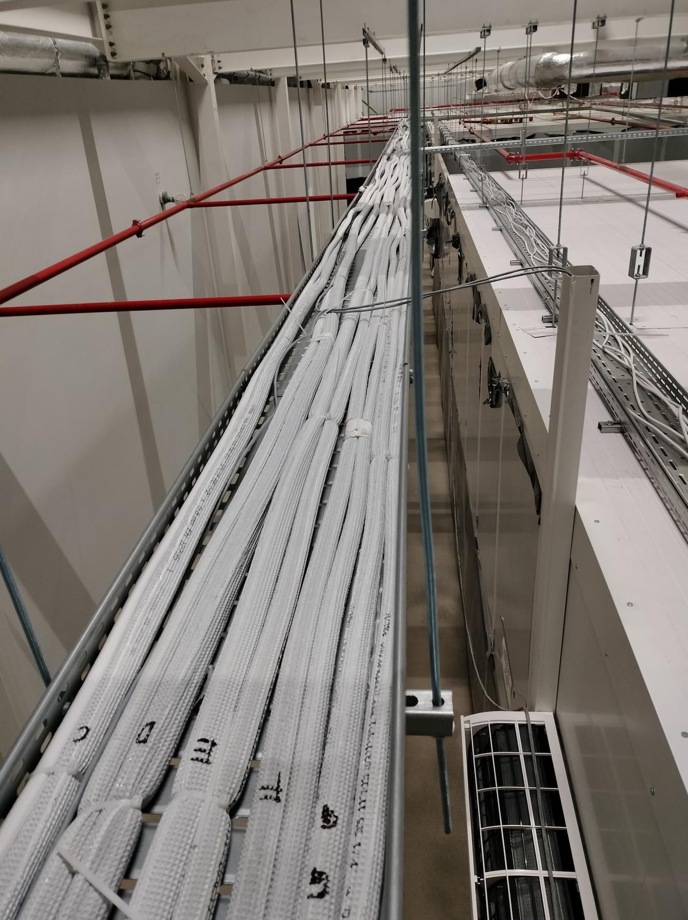 Białe, zaizolowane rury instalacyjne ułożone w metalowej korycie pod sufitem, widoczne czerwone rury i fragment klimatyzatora, w tle białe ściany hali.