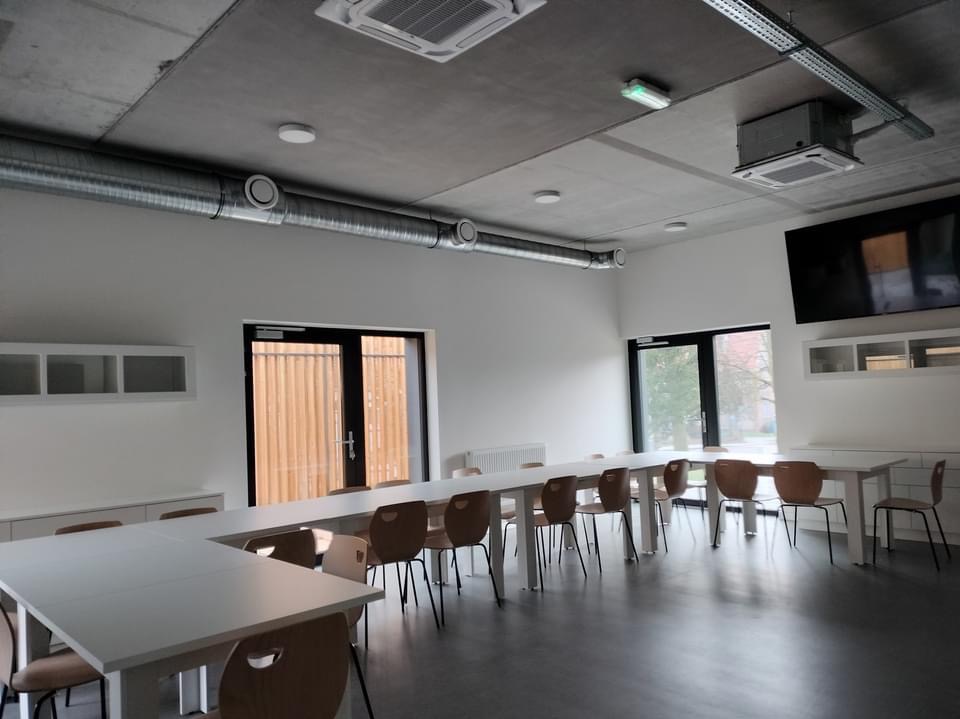 Wnętrze biura z systemem wentylacji: widoczne kanały wentylacyjne na suficie, kratki wentylacyjne w suficie podwieszanym, białe ściany, długi stół konferencyjny z krzesłami, szare panele podłogowe.