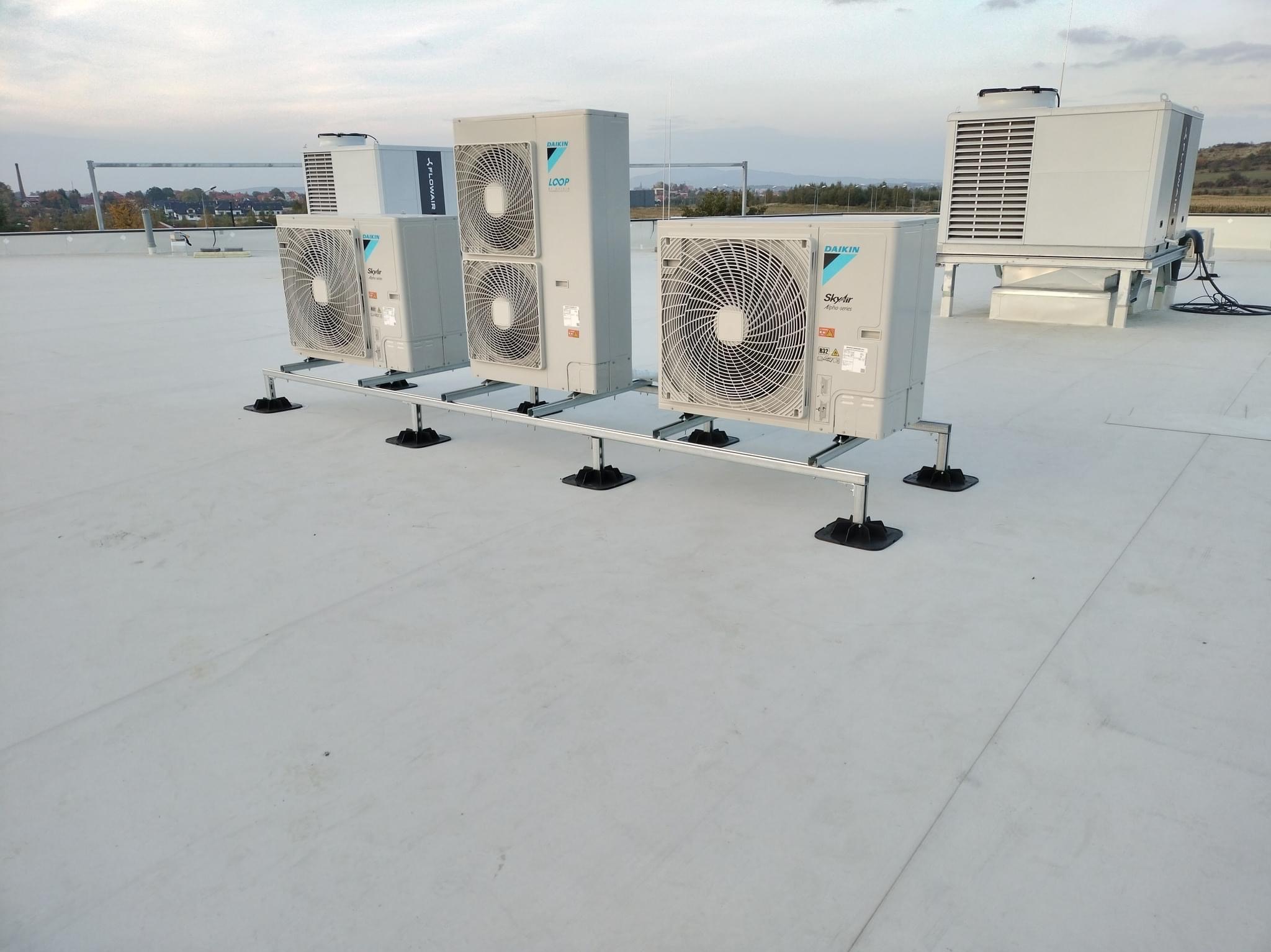 Instalacja kilku jednostek zewnętrznych klimatyzacji Daikin na płaskim dachu, z widocznymi metalowymi konstrukcjami wsporczymi i czarnymi podkładkami antywibracyjnymi, w tle krajobraz miejski...