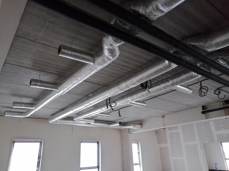 Widok instalacji wentylacyjnej z aluminiowymi kanałami pod sufitem w surowym wnętrzu budynku, z widocznymi przewodami i fragmentami ścian przygotowanych do malowania.