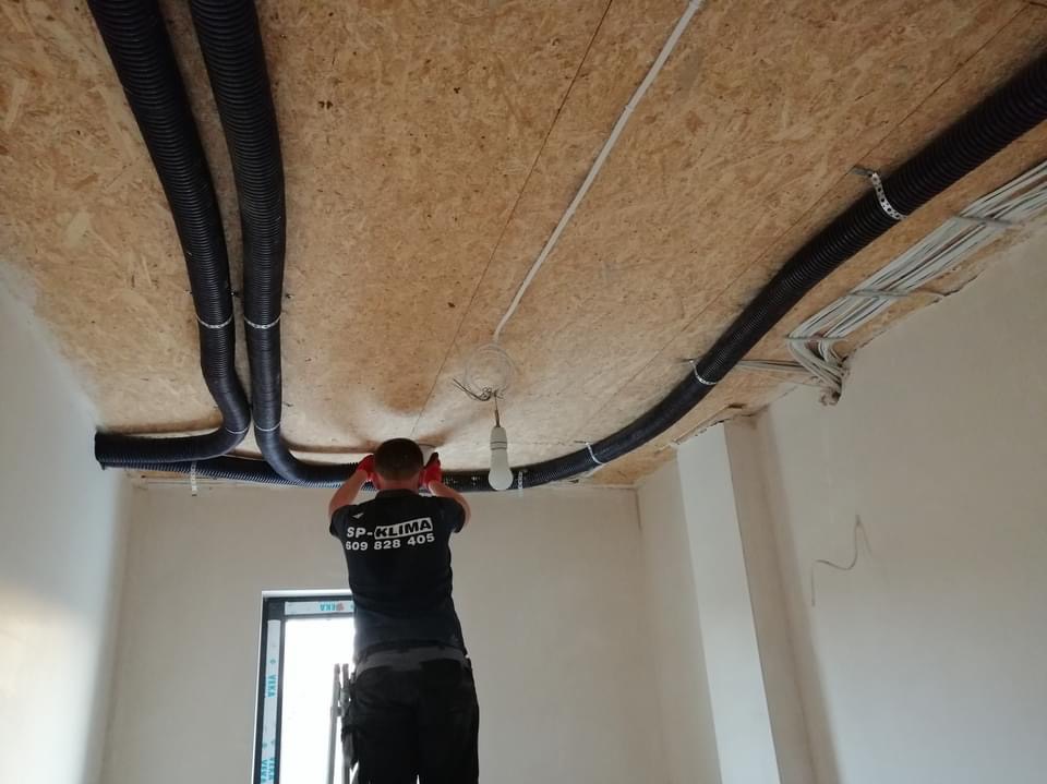 Montaż czarnych, karbowanych rur wentylacyjnych pod sufit z płyty OSB w nowym budynku, widoczny pracownik w czerwonych rękawicach instalujący system, zwisająca żarówka i wystające kable.